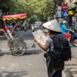 Viajera con sombrero cónico, mochila y mapa caminando por una calle del Old Quarter de Hanoi entre motos y un cyclo