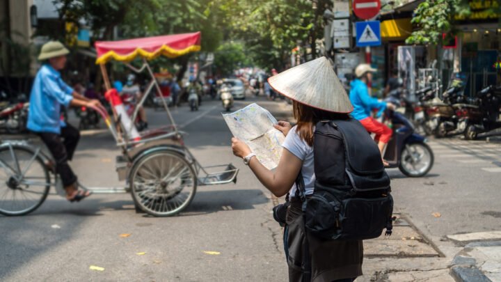 Viajera con sombrero cónico, mochila y mapa caminando por una calle del Old Quarter de Hanoi entre motos y un cyclo