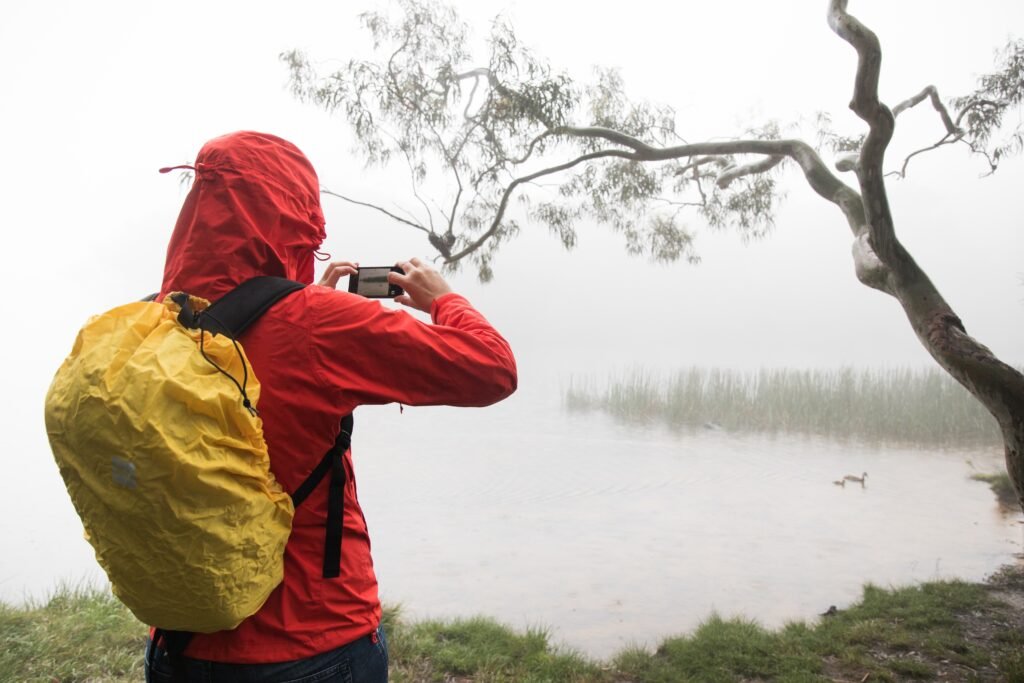 Viajera con chubasquero rojo y funda impermeable amarilla en la mochila haciendo una foto junto a un lago