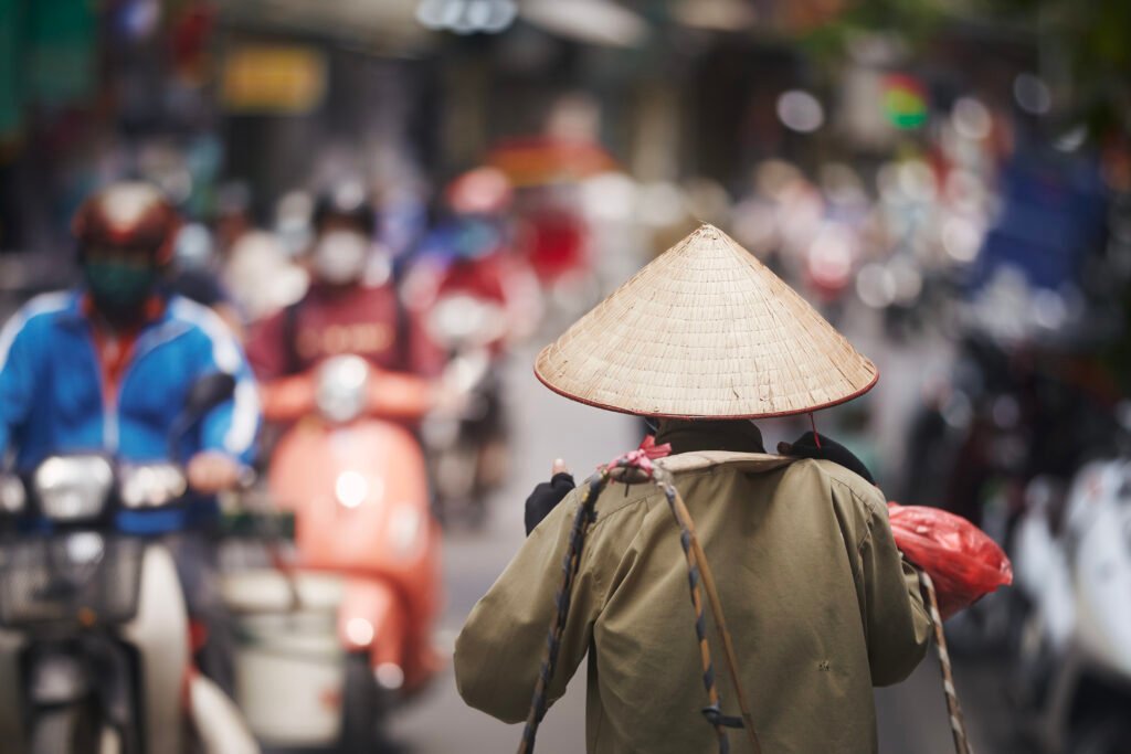 Vendedora ambulante con sombrero cónico y pértiga de bambú al hombro caminando entre el tráfico del Old Quarter de Hanoi