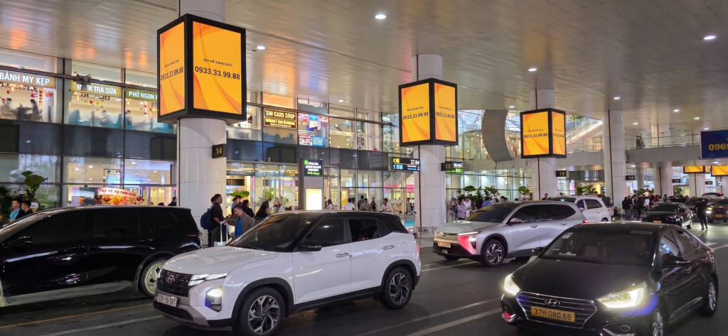 Zona exterior de llegadas en un aeropuerto de Noi Bai (Hanoi), con pasajeros esperando en la acera, coches recogiendo viajeros y grandes columnas con pantallas publicitarias amarillas.