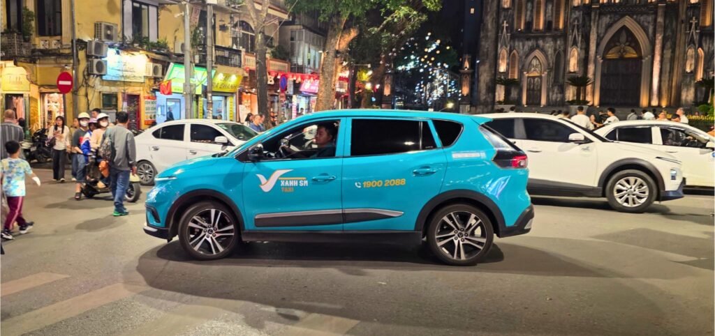 Taxi eléctrico cian de la empresa Green SM navegando por el tráfico nocturno de Hanoi, con la Catedral de San José de fondo.
