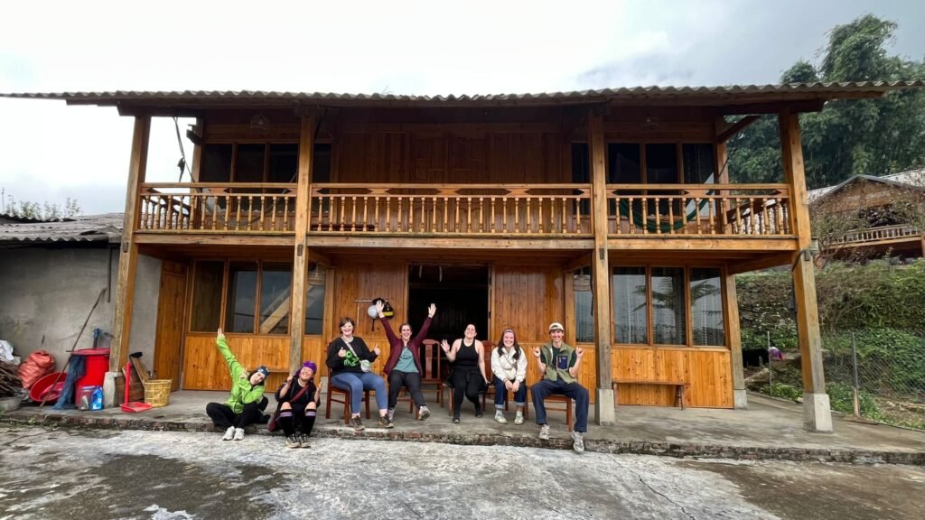 Casa de madera de dos plantas de Mama Vu Vi con un grupo de viajeros sentados en la entrada, valle de Sapa