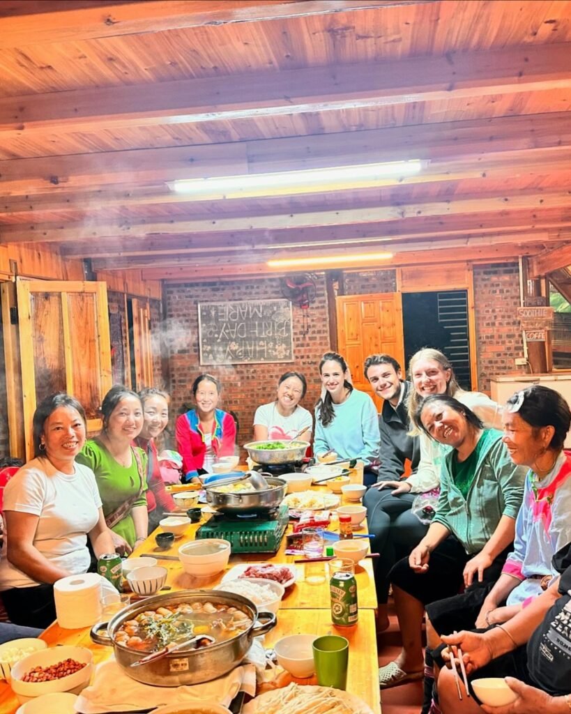 Viajeros y mujeres hmong cenando hotpot en una mesa larga en el alojamiento de Dinh Sapa Trekking Adventures