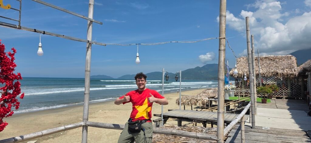 Viajero en un restaurante de bambú sobre la playa de Lang Co, con el mar y las montañas al fondo, durante una parada del Hai Van Pass