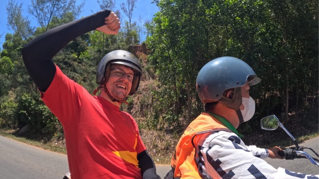 Alberto sonriente con el brazo en alto detrás de un conductor local de easyrider durante el tour del Hai Van Pass